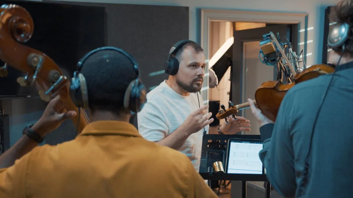 Tar Sahno in headphones, directing violin and bass in the studio, a score open on the stand